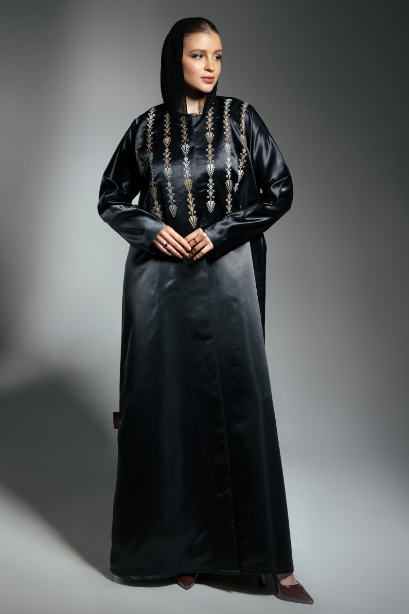 A woman wearing a traditional black abaya with vertical embroidered motifs in gold and cream tones, paired with a matching headscarf.