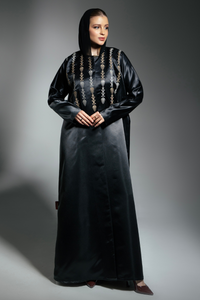 A woman wearing a traditional black abaya with vertical embroidered motifs in gold and cream tones, paired with a matching headscarf.